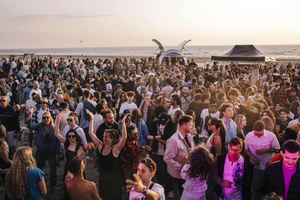 Grote groep mensen danst en feest op het strand tijdens zonsondergang, met de zee op de achtergrond en warme avondzon die over de menigte schijnt.
