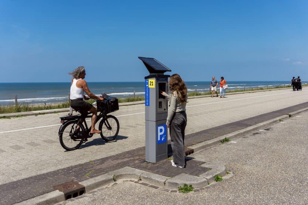 Persoon gebruikt een parkeerautomaat op de boulevard van Zandvoort, terwijl een fietser passeert met uitzicht op strand en zee.