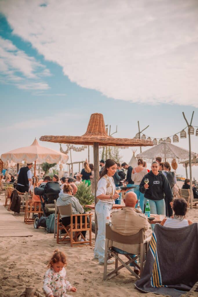 Bezoekers genieten van eten en drinken op een sfeervol strandterras met parasols en zitplaatsen in het zand.