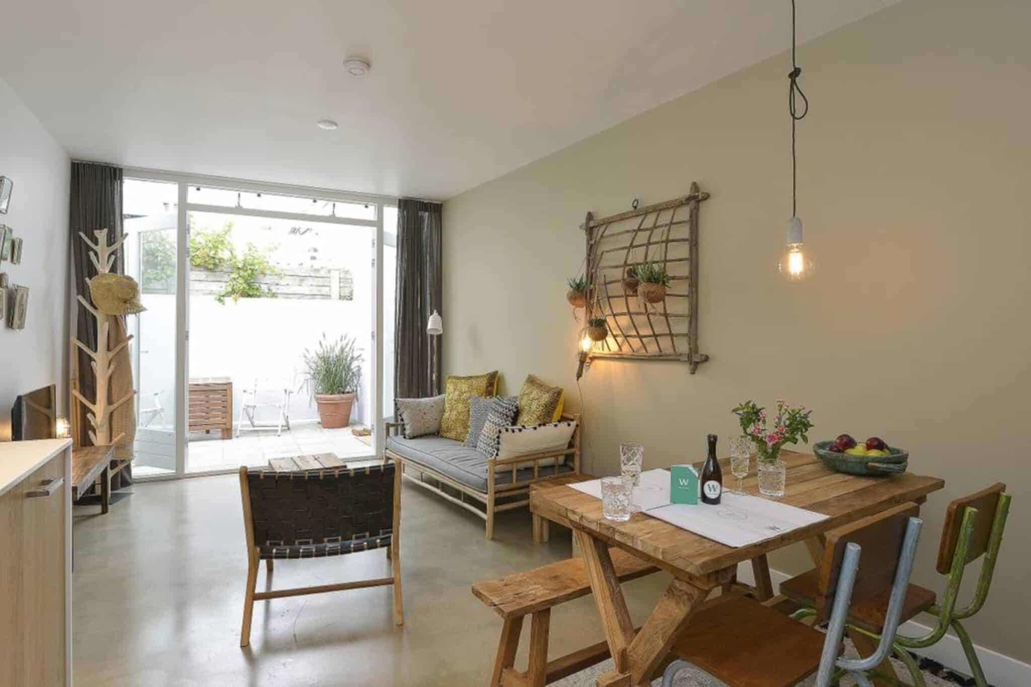 Lichte woonkamer van appartement Folloew the sun in centrum van Zandvoort met houten eettafel, bank met kussens, wanddecoratie en openslaande deuren naar buiten.