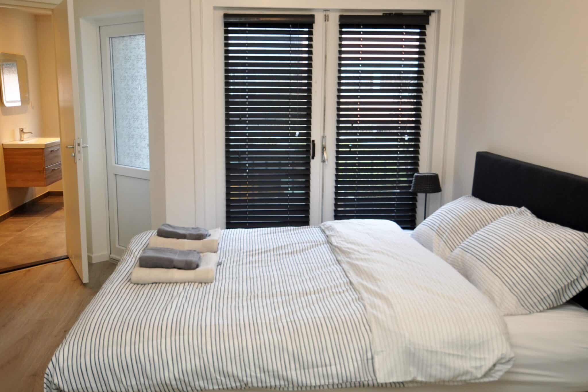 Lichte moderne slaapkamer met tweepersoonsbed, zwart hoofdbord, jaloezieën en open badkamer met houten wastafel.
